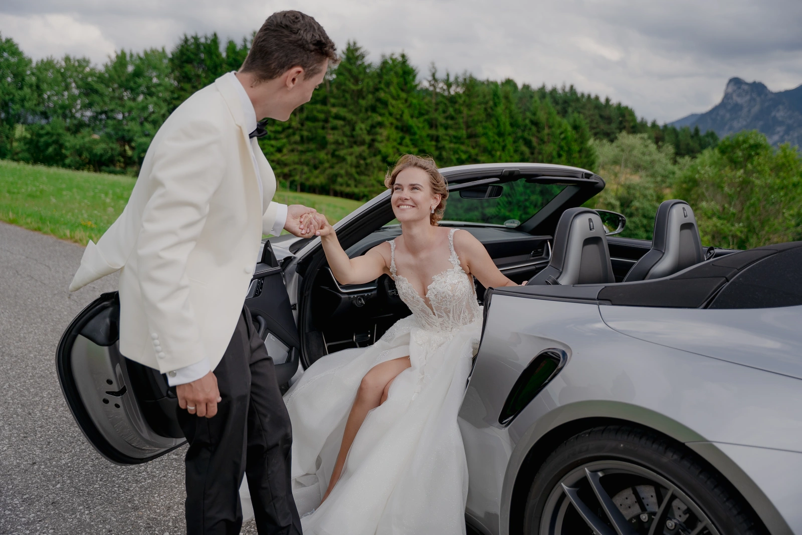Braut steigt bei einem Hochzeitsshooting im Allgäu aus einem Porsche, während der Bräutigam ihr die Hand reicht.
