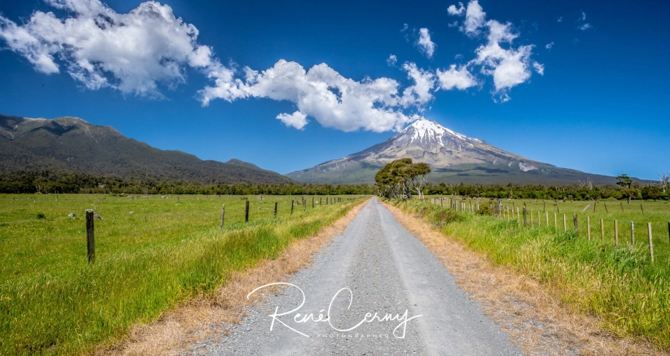 Fotograf René Cerny – Landschaftsfotografie. Schotterstraße durch grünes Farmland mit Blick auf den Vulkan Taranaki, Neuseeland.