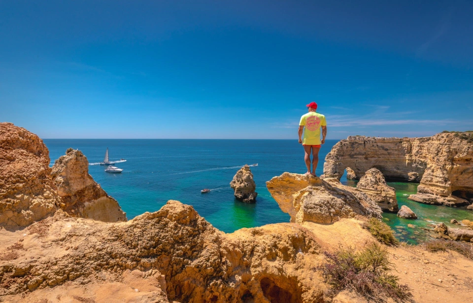 Fotograf René Cerny – Reisefotografie. René steht auf einer Felsklippe über einer Bucht mit Segelbooten, Algarve, Portugal.