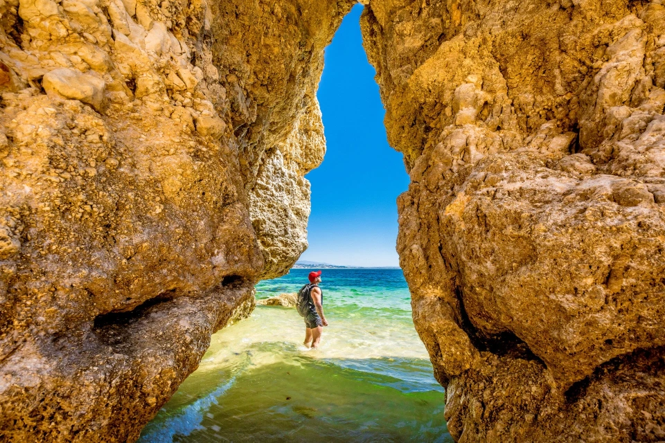 Fotograf René Cerny – Reisefotografie. René watet durch türkisfarbenes Wasser zwischen zwei Felsentoren, Algarve, Portugal.