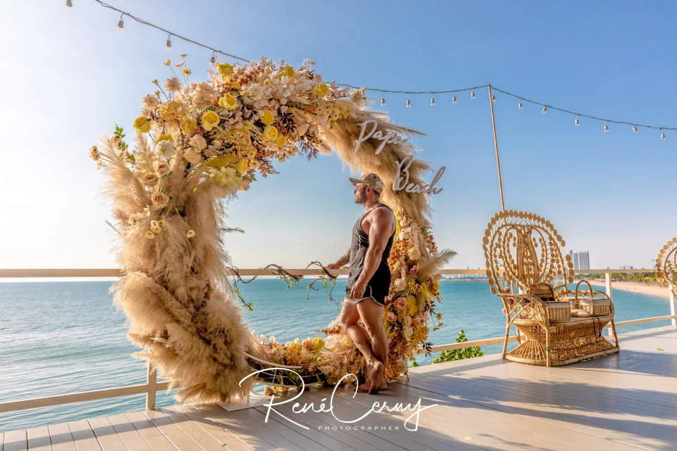 Fotograf René Cerny – Reisefotografie. René steht am Strand vor einem Strand-Schild mit Blumenbogen, Thailand.