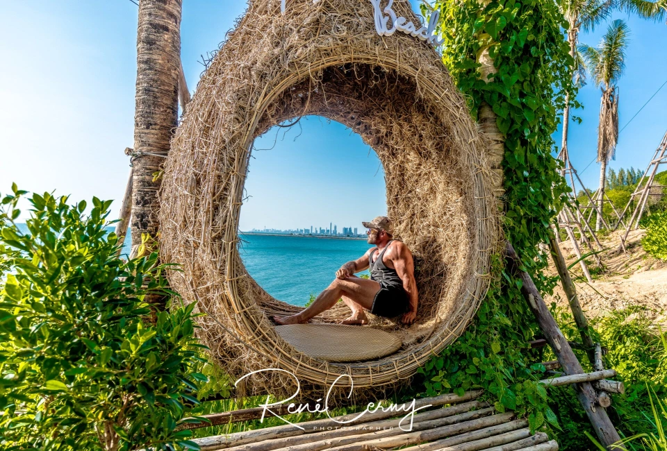 Fotograf René Cerny – Reisefotografie. René sitzt in einer geflochtenen Strandschaukel mit Blick auf Meer und Skyline, Thailand.
