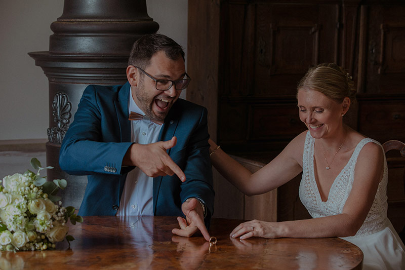 Hochzeitspaar sitzt am Tisch und Bräutigam zeig mit dem Finger auf die Ringe. Der Fotograf aus München fotografiert, Hochzeitsfotos, Hochzeitsfotograf Preise Augsburg.