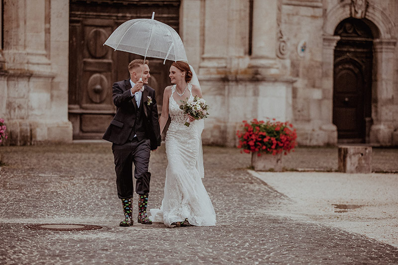 Hochzeitspaar beim Regen mit Regenschirm und Gummistiefel. Hochzeitsfotograf München begleitet sie, Hochzeitsfotos für die Galerie