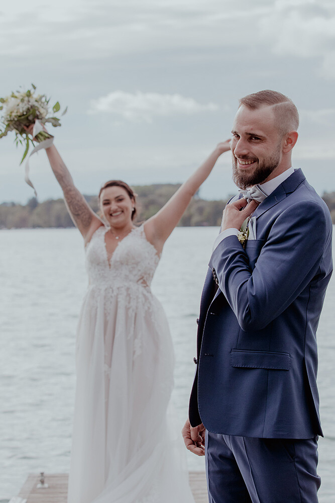Gutes Wetter Garantie. Bräutigam posiert vor der Kamera am See und die Braut winkt ihm zu im Hintergrund. Hochzeitsfotograf Preise Augsburg.