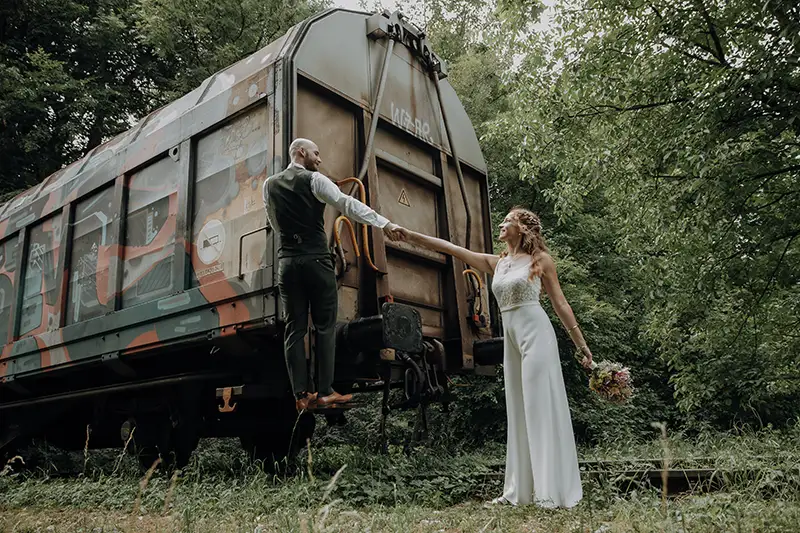 Braut und Bräutigam halten sich an einer mit Graffiti bedeckten Schleppe in einer üppigen, grünen Umgebung an den Händen.