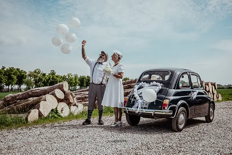 Ein älteres Paar in Hochzeitskleidung lässt neben einem geschmückten Oldtimer und gestapelten Holzscheiten im Freien Luftballons steigen.